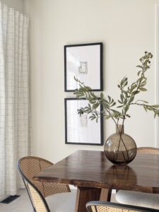 Expertly designed room with white finishes. Brown wood table with round vase and greenery. Two black and white images on the wall behind table. 